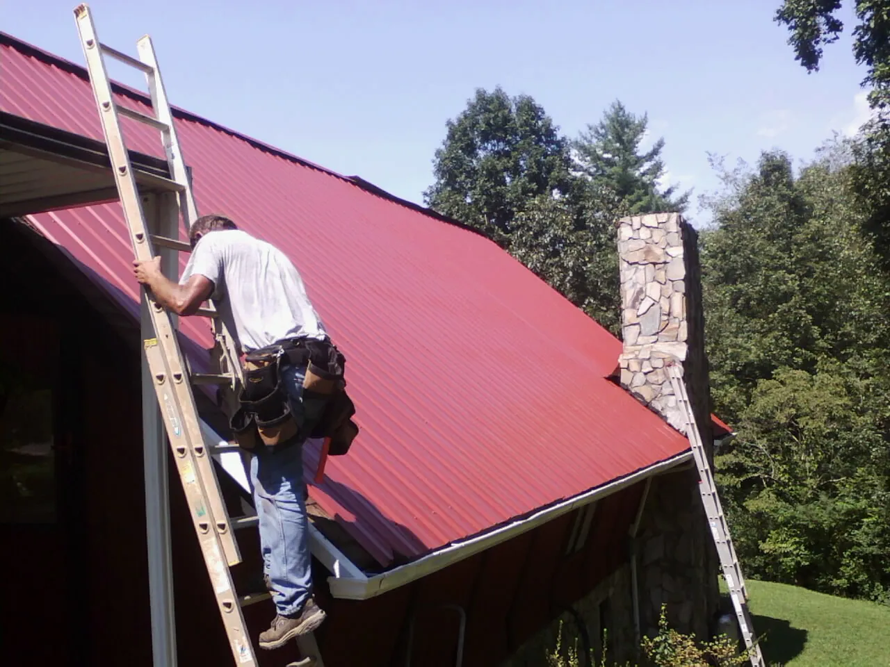 Roofer on a ladder beside a red metal roof and stone chimney — Soffit & Fascia Repair by Roofing Brothers in Plover